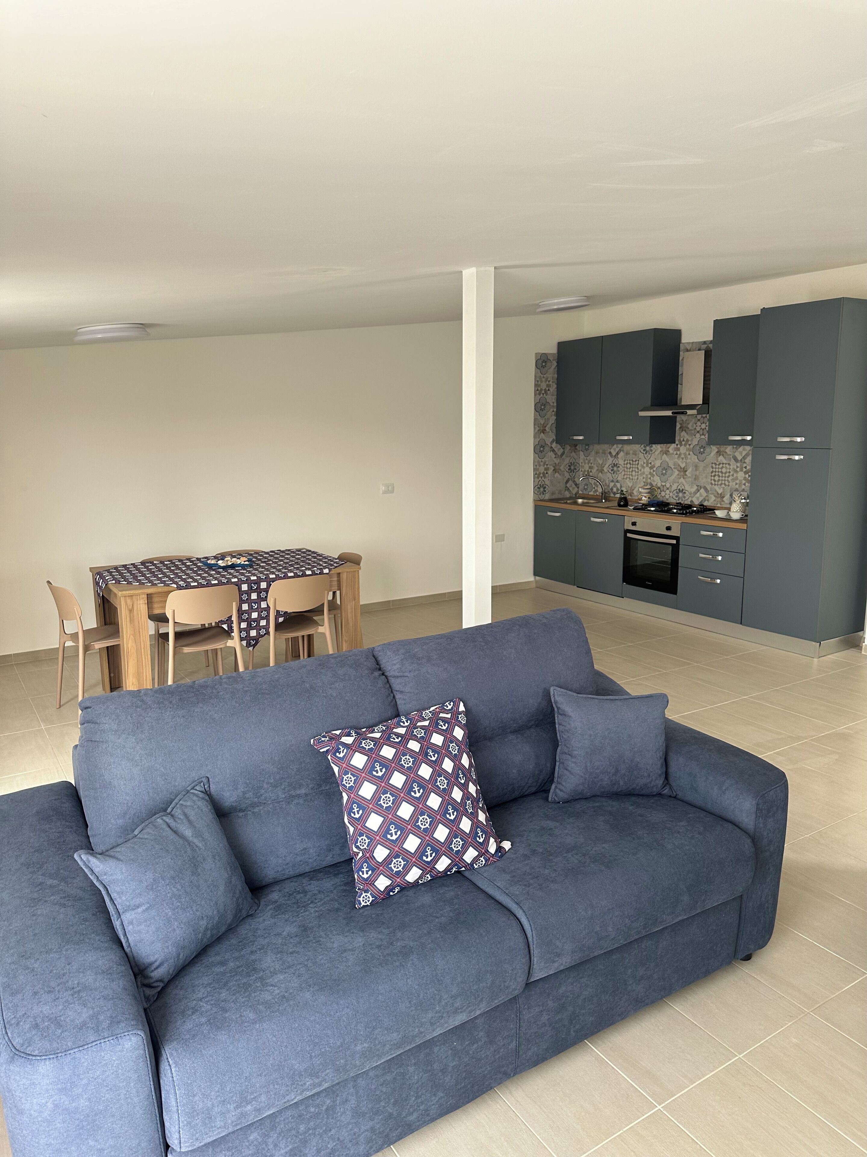 A modern living room with a blue sofa and patterned cushions, a dining area with a wooden table and chairs, and a kitchen with grey cabinets and a tiled backsplash.