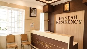 Un espace d’accueil comprenant un comptoir de réception portant l’inscription « GANESH RESIDENCY », deux chaises, un tableau encadré accroché au mur et une fenêtre laissant entrer la lumière naturelle.