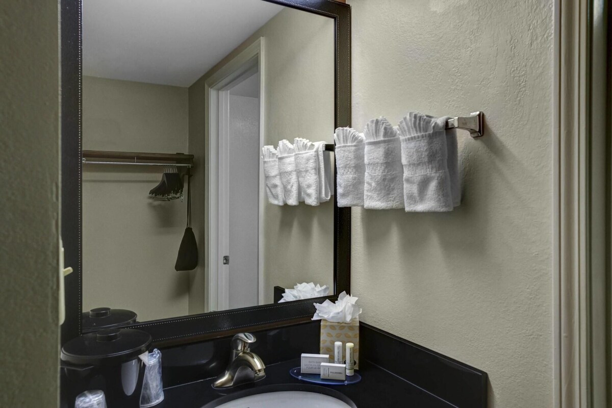A bathroom with a large mirror, a towel rack with multiple towels, a sink with toiletries, and a door leading to a shower area.