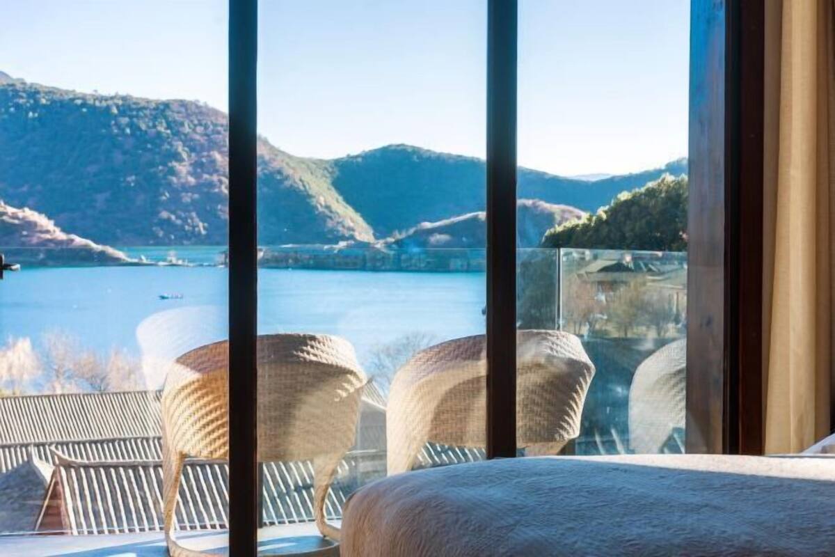 Een kamer met een groot raam met uitzicht op een waterpartij en heuvels. Er staan rieten stoelen en een bed.