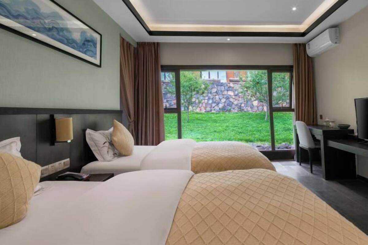 Hotelkamer met twee bedden, een bureau, een televisie en een groot raam met uitzicht op groen en een stenen muur.