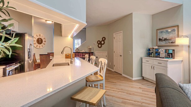 Una cocina moderna con una isla central, una zona de comedor con sillas, un sofá y un reloj de pared.