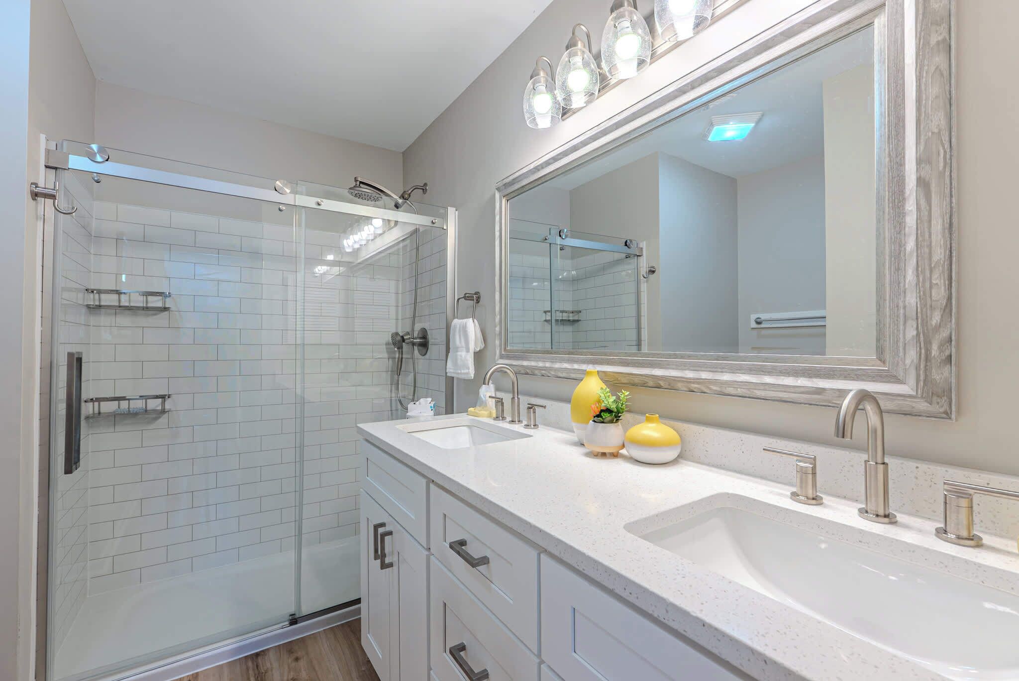 Une salle de bain moderne dotée d’une douche à parois de verre, d’un grand miroir et d’un plan de toilette blanc avec deux vasques.