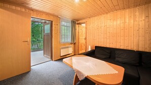 Interior de una cabaña de madera con un sofá, una mesa cubierta con un mantel blanco, una ventana con vistas a la vegetación y una puerta que da al exterior.
