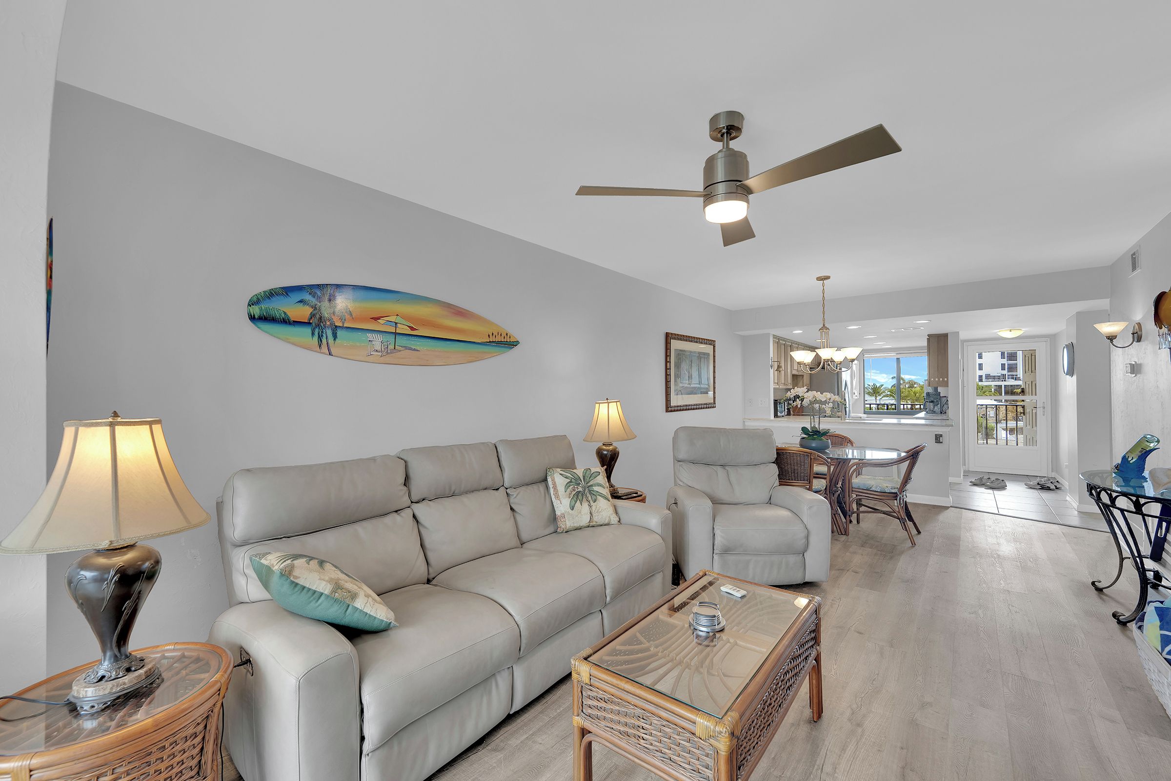 Un salon comprenant un canapé, une table basse, une lampe, un ventilateur de plafond et un tableau représentant une scène de plage.