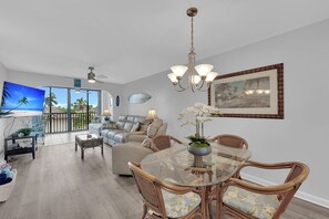 A spacious living area with a large flat-screen TV, comfortable seating, a dining table with chairs, and a balcony with a view. - SUMMER SPECIAL DISCOUNT!!!  2B/2B Fairview Isles 214 Condo, Walk to Beach, Heated Pool! (Fort Myers Beach)