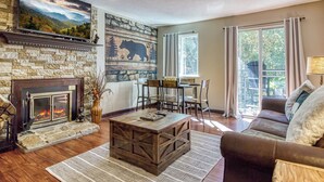 Una sala de estar con chimenea, pared de piedra y televisor grande. Hay sillones y un sofá, y una ventana con cortinas.
