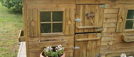 A wooden cabin with a sloped roof, a large window, and a door with a small window. There is a planter with flowers and two small figurines on the ground.