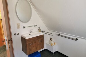 A small bathroom with a white sink, wooden vanity, and a mirror. There are grab bars on the walls and a blue trash bin. - Vacation apartment Mary (Karlsburg)