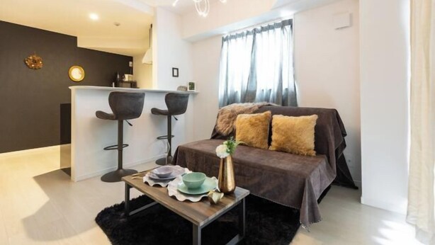 A modern living room with a sofa, two bar stools, a coffee table, and a small kitchen area. - Ra Premier K (Osaka)