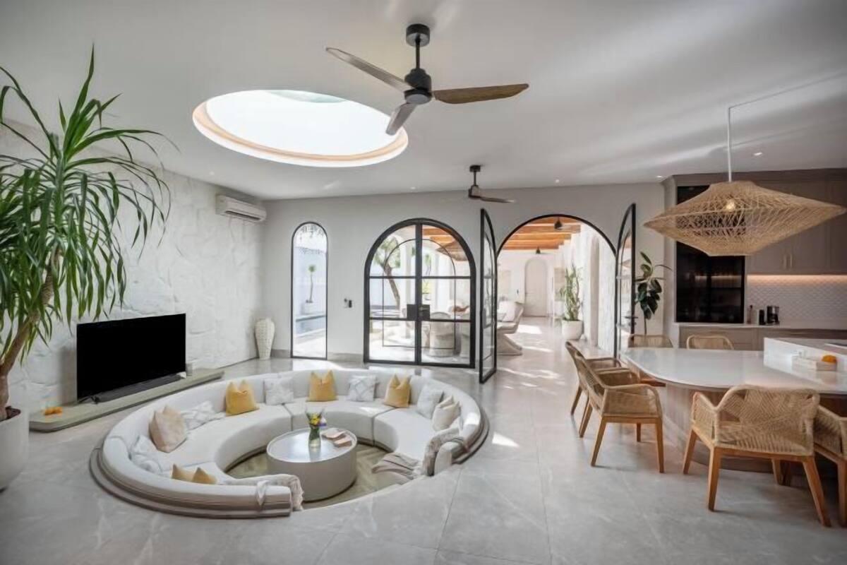 A modern living room with a curved sofa, a flat-screen TV, and a dining area with wicker chairs.