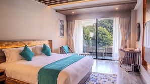 A large bed with a wooden headboard, a teal throw blanket, and two teal pillows. There is a small table and a chair near the window, and a balcony with a view of greenery. - Soaltee Westend Lakeside Pokhara (Pokhara)