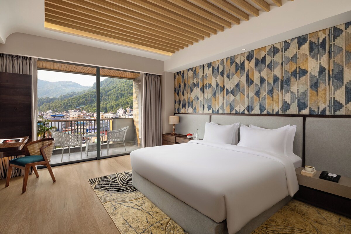 Habitación de hotel con una cama grande, un escritorio y un balcón con vistas a la ciudad y las montañas.