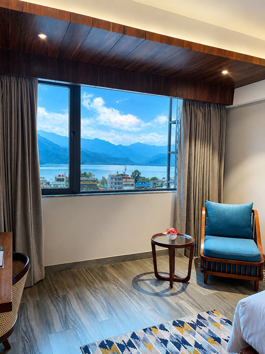 Ein Hotelzimmer mit großem Fenster, das Blick auf einen See und Berge bietet, einer Holzzieldecke, einem blauen gepolsterten Sessel, einem kleinen runden Tisch mit einer Vase und einem gemusterten Teppich.