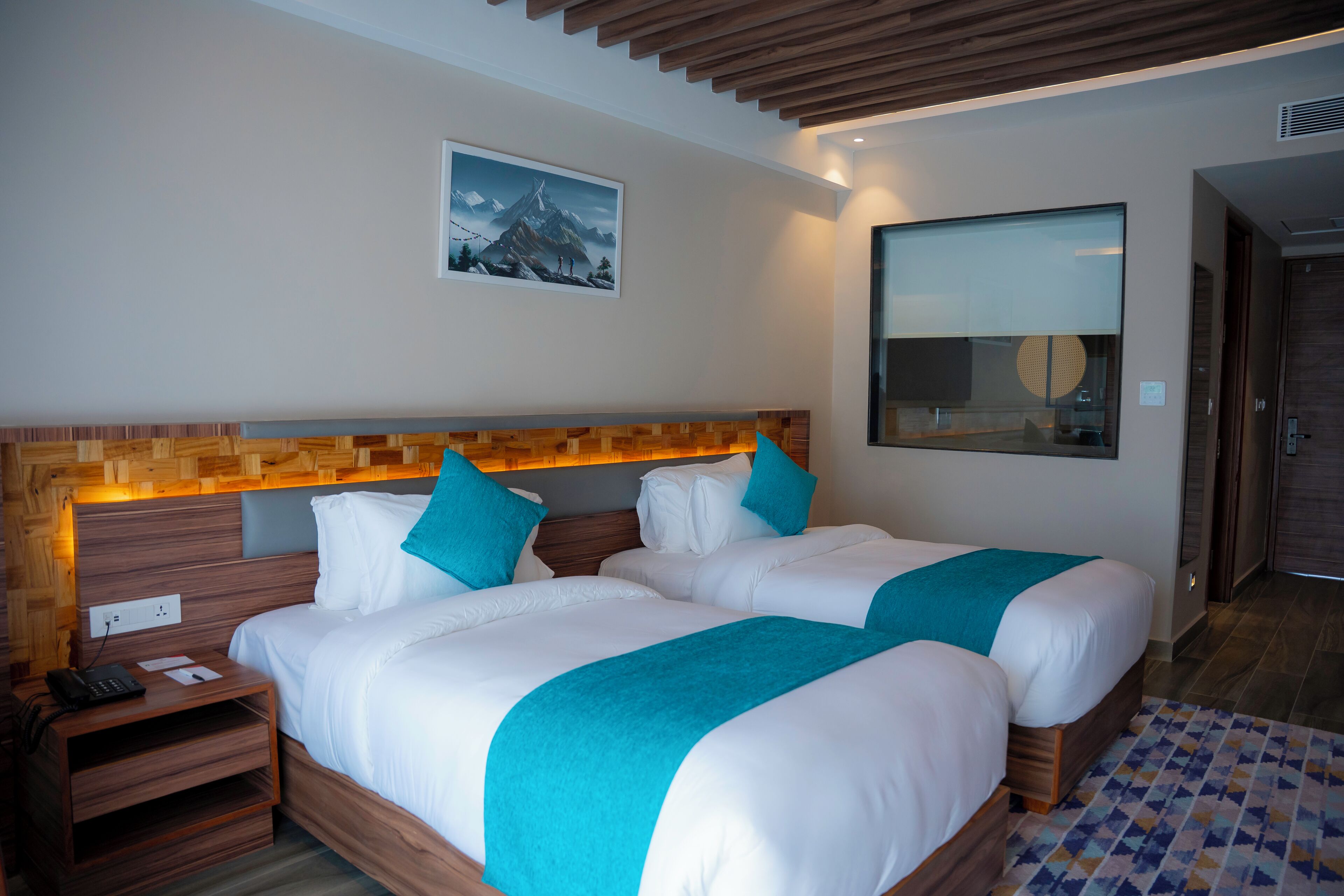 A hotel room with two beds, wooden headboard, a painting, and a large window.