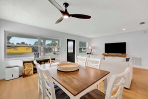 Una sala moderna con una mesa de comedor de madera, sillas blancas, un ventilador de techo y un amplio ventanal con vista a una casa amarilla.