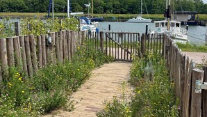 A wooden pathway leading to a dock with boats, surrounded by greenery and a clear sky.