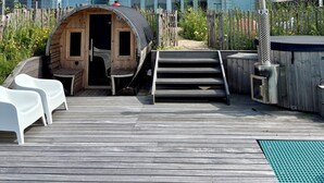 A wooden deck with steps leading to a small wooden structure, possibly a sauna or hot tub, surrounded by a wooden fence and a row of houses with solar panels.
