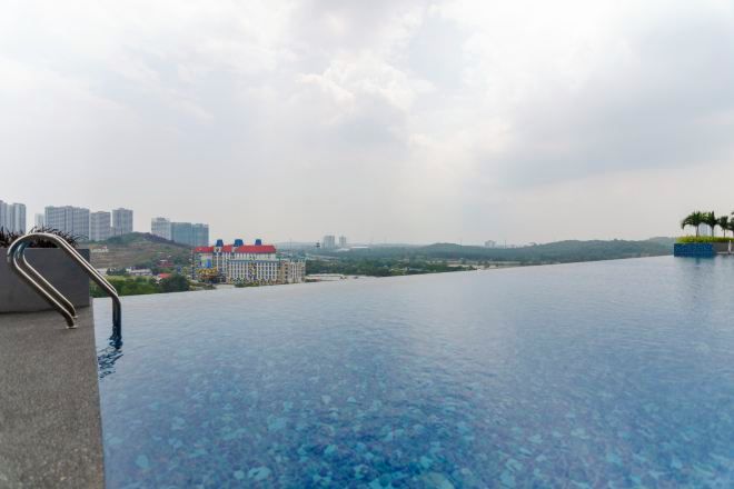 A clear view of an infinity pool with a metal handrail on the left, a red-roofed building in the background, and a distant cityscape.