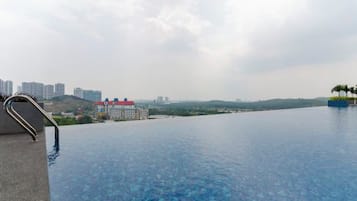 A clear view of an infinity pool with a metal handrail on the left, a red-roofed building in the background, and a distant cityscape.