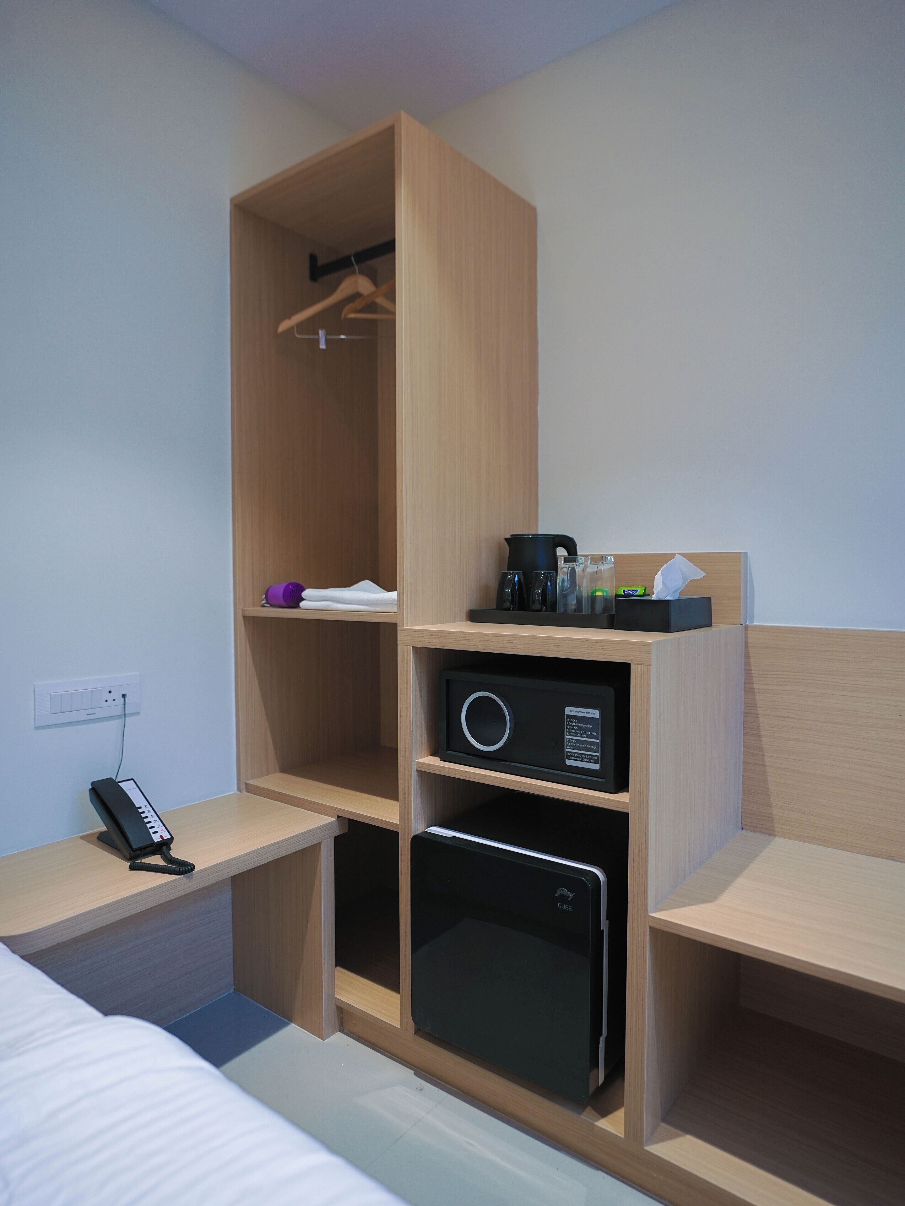 A hotel room with a wooden wardrobe, a bedside table with a phone, a small black appliance, and a coffee maker.