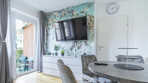 A modern living room with a flat-screen TV, a dining table with chairs, a sofa, and a view through a glass door to an outdoor area with chairs. - Cozy 1BR+1LR w/ Balcony, 9-Min to City Center (Vienna)