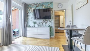 A modern living room with a flat-screen TV, a white console, a dining table with chairs, and a view through a sliding glass door. - Cozy 1BR+1LR w/ Balcony, 9-Min to City Center (Vienna)