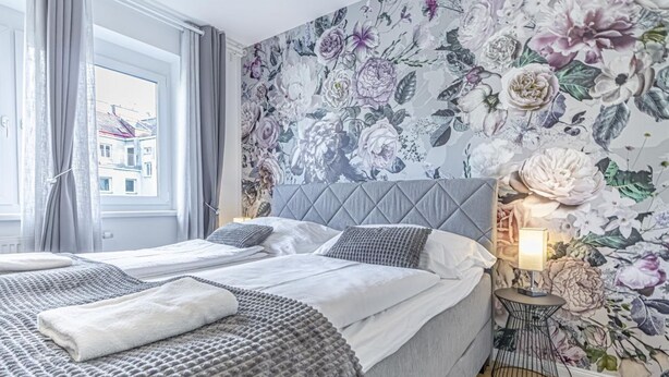 A bedroom with a bed, pillows, a bedside table, a lamp, and a floral wallpaper. - Cozy 1BR+1LR w/ Balcony, 9-Min to City Center (Vienna)