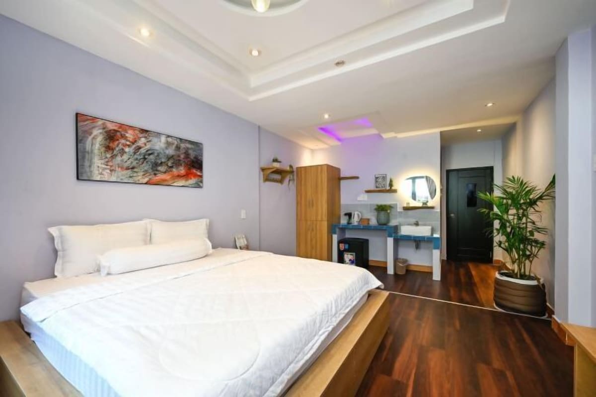 Une chambre moderne avec un grand lit, un plancher en bois, un tableau au mur et une petite kitchenette à l’arrière-plan.