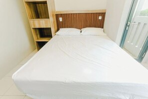 Una cama individual con cabecera de madera, una pequeña estantería de madera y un ventanal corredero.