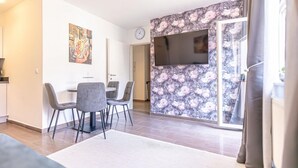 Ett modernt vardagsrum med en platt-TV som är monterad på en vägg med ett blommönster, en matplats med ett bord och stolar samt ett stort fönster med gardiner.
