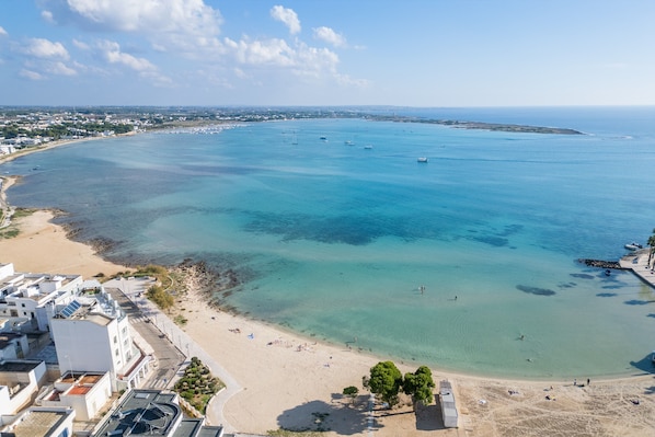 An aerial view of a coastal area with a beach, clear blue waters, and a distant shoreline with buildings.