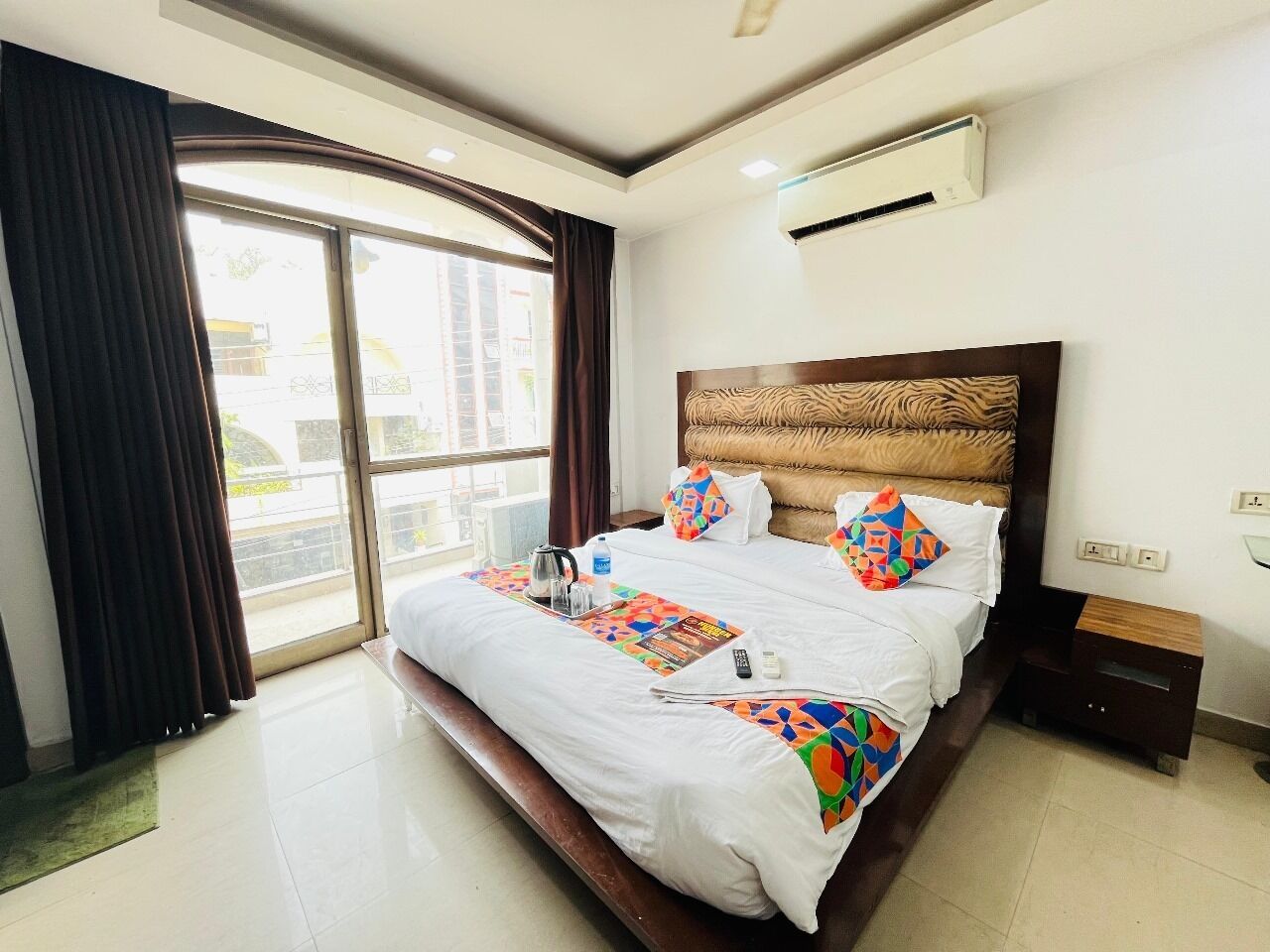 A hotel room with a large bed, a bedside table, an air conditioning unit, and a view through a large window.