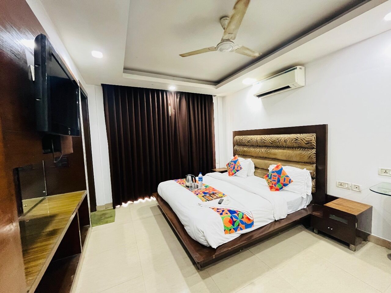 A hotel room with a bed, a TV, a ceiling fan, and an air conditioning unit.