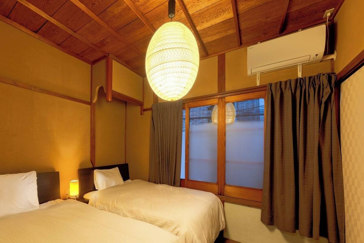 A hotel room with two beds, wooden ceiling beams, a large window with curtains, and a hanging light fixture.