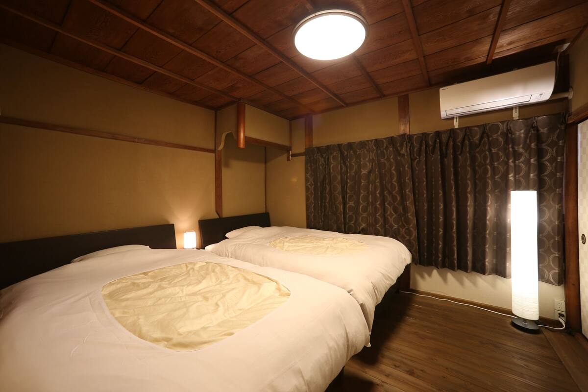 A room with two beds, wooden ceiling beams, an air conditioning unit, a window with curtains, and a wall-mounted light.