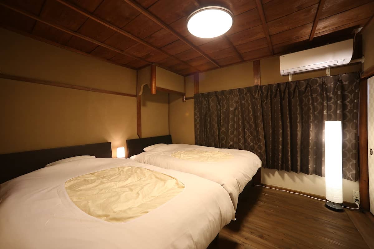 A room with two beds, wooden ceiling beams, an air conditioning unit, a window with curtains, and a wall-mounted light.