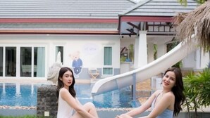 Two people sitting by a pool with a slide, a house with a red roof, and a stone wall in the background. - Destiny Pool Villa (Bang Lamung)