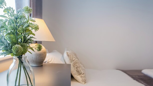A neatly arranged room with a bed, a bedside lamp, a vase with green plants, and a small decorative item on the nightstand. - 9 Monks Crescent (Durham)