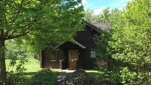 A rustic log cabin with a wooden door and a small window, surrounded by lush greenery and a clear sky.