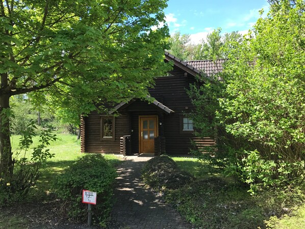 A rustic log cabin with a wooden door and a small window, surrounded by lush greenery and a clear sky.