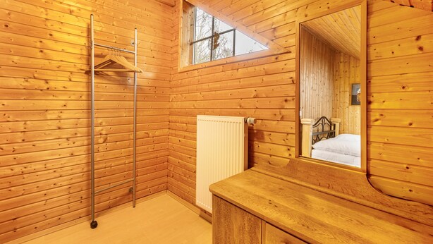 A wooden cabin interior with a wooden floor, a radiator, a wooden dresser, a mirror, and a window with a view of trees.