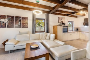 Un salon moderne avec un canapé d’angle, une table basse en bois, un coin cuisine équipé d’appareils électroménagers et des œuvres d’art accrochées aux murs.