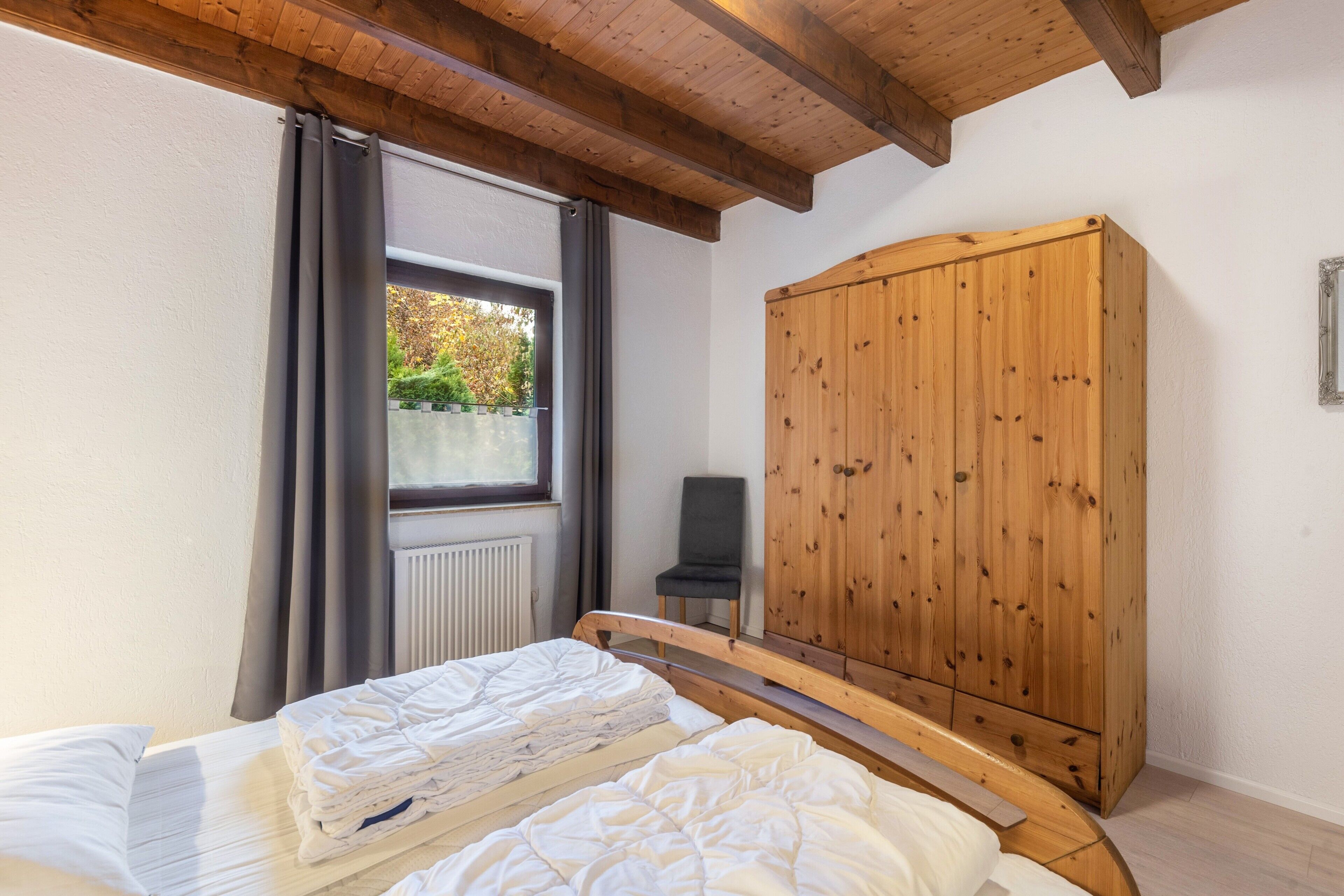 Un dormitorio con cama, armario de madera, una silla y una ventana con cortinas.