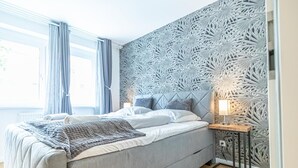Et moderne soveværelse med en seng, et natbord, en væg med geometrisk mønster og et vindue med gardiner.