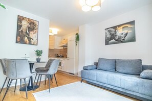 Moderni olohuone, jossa on harmaa sohva, ruokapöytä ja tuolit. Taustalla näkyy keittiö, ja seinillä on kaksi luontoaiheista taideteosta.