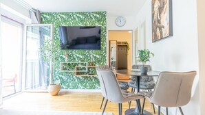 Et moderne spiseområde med et stort bord, grå polstrede stole og et vægmonteret tv.