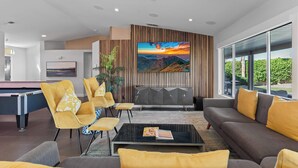 Ein modernes Wohnzimmer mit Billardtisch, gelben Sesseln, einem grauen Sofa und einem großen Fernseher an der Wand.