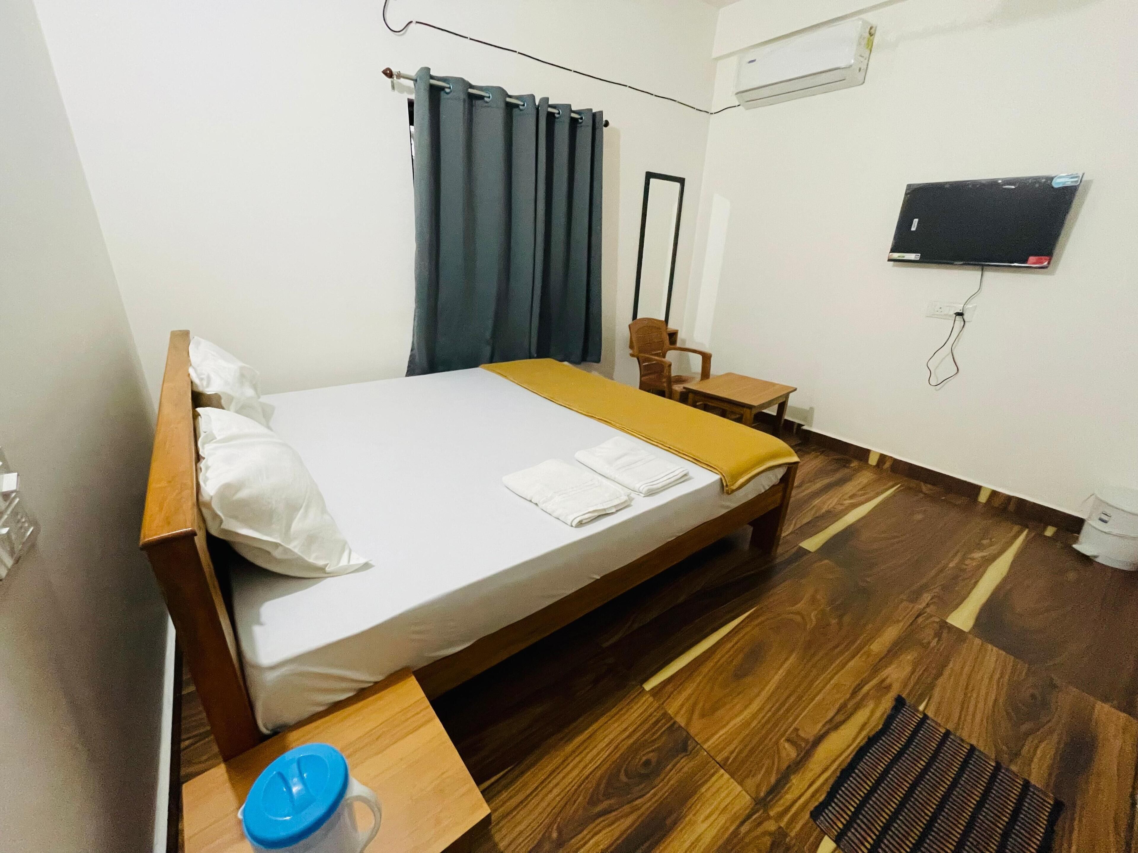 Een opgemaakt bed met houten hoofdeinde, een klein nachtkastje met een blauwe beker, een stoel, een bureau en een aan de muur bevestigde televisie.