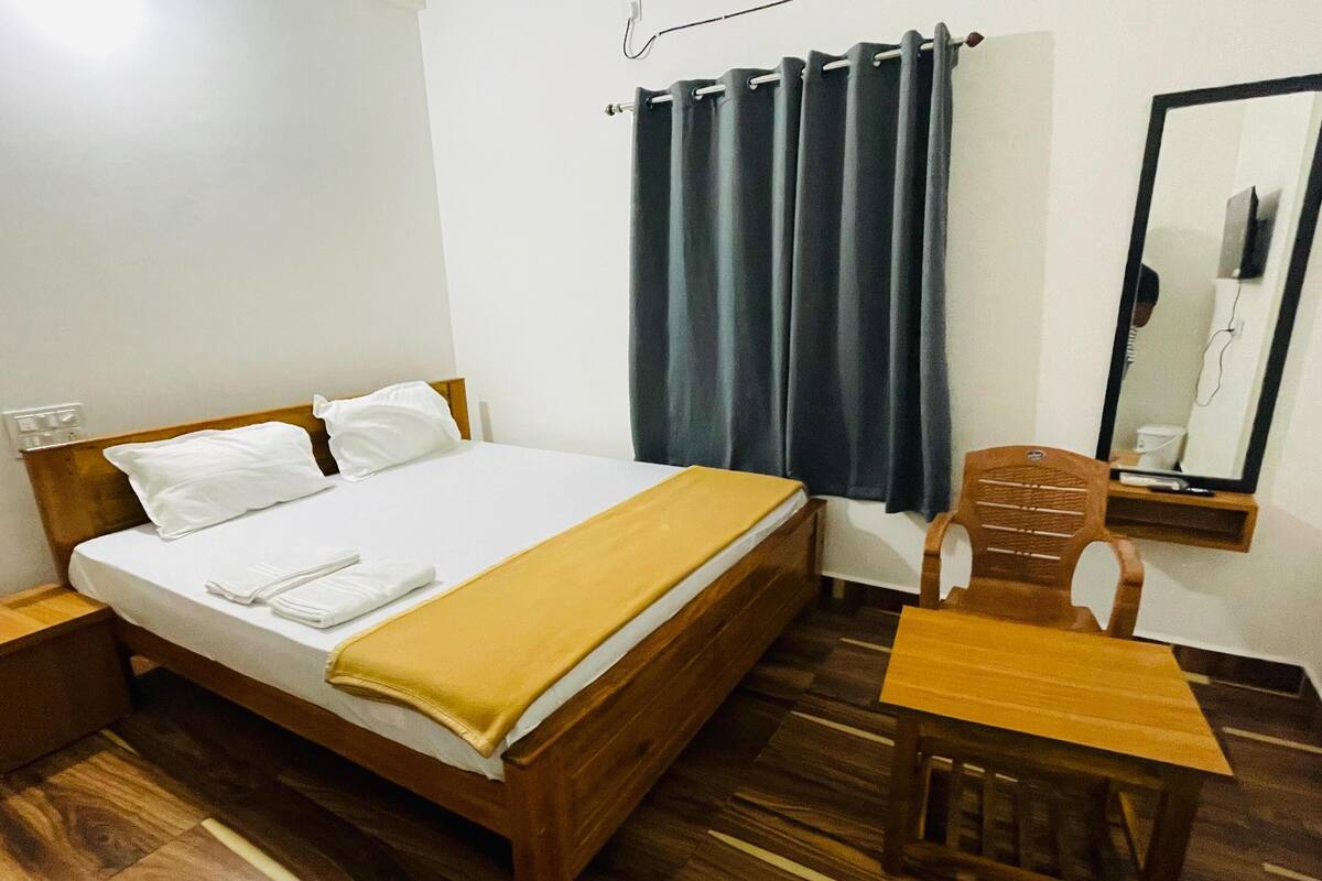 A single bed with a wooden headboard, white pillows, and a yellow blanket. There is a wooden chair and a small table beside the bed. A dark curtain hangs on the window, and a mirror is mounted on the wall.